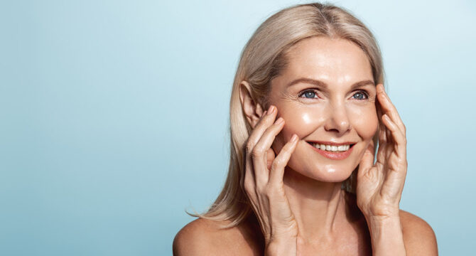 Smiling middle aged woman, 50 years model, applies anti-aging skin cream, rubbing in moisturizer on face, standing over blue background