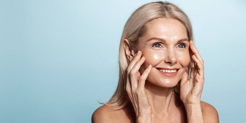 Smiling middle aged woman, 50 years model, applies anti-aging skin cream, rubbing in moisturizer on face, standing over blue background
